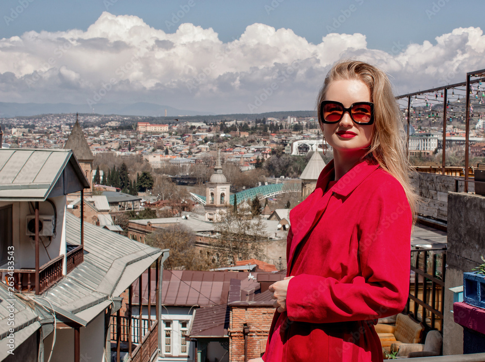 A beautiful young girl blonde in sunglasses and a red coat, stands on the background of the city of Tbilisi. Georgia.