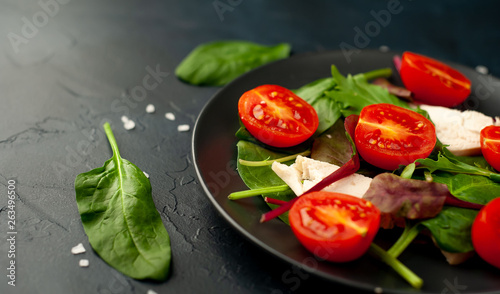Mix fresh leaves of arugula, lettuce, spinach, tomatoes and chicken fillet for salad, on a dark plate against a background of concrete
