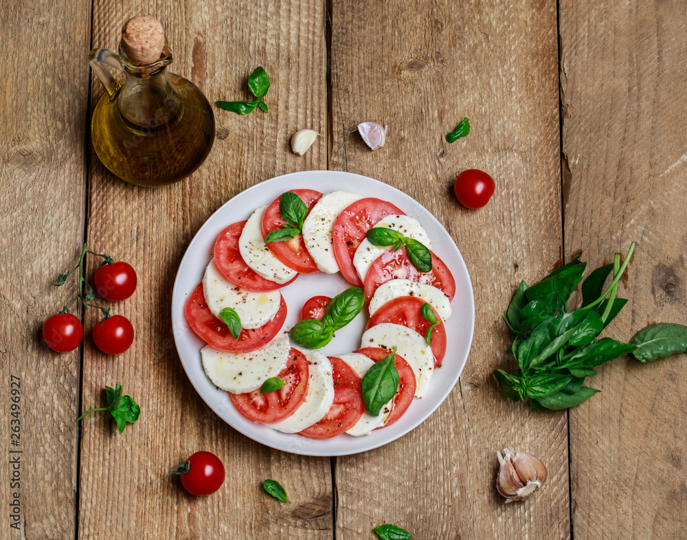 Delicious italian caprese salad with ripe tomatoes, fresh basil and mozzarella cheese on wooden rustic background. Italian caprese salad in white plate with sliced tomatoes.