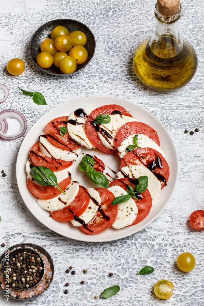 Italian caprese salad with ripe tomatoes, fresh basil and mozzarella cheese on grey stone background. Italian caprese salad in white plate with sliced tomatoes