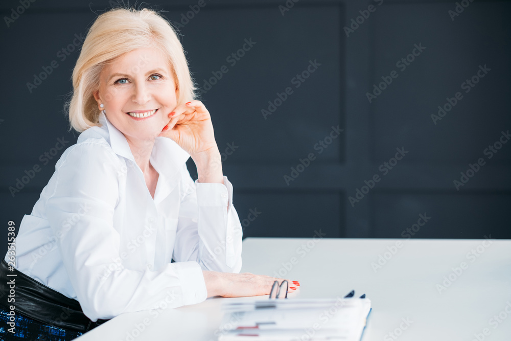 Fototapeta premium Smiling business woman at workplace. Time for break. Aged lady sitting looking at camera. Hand at cheek. Copy space.