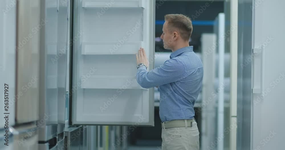 The man open the door of the refrigerator in the appliances store and compare with other models to buy the new house The man open the door of the refrigerator in the appliances store and compare with other models to buy the new house