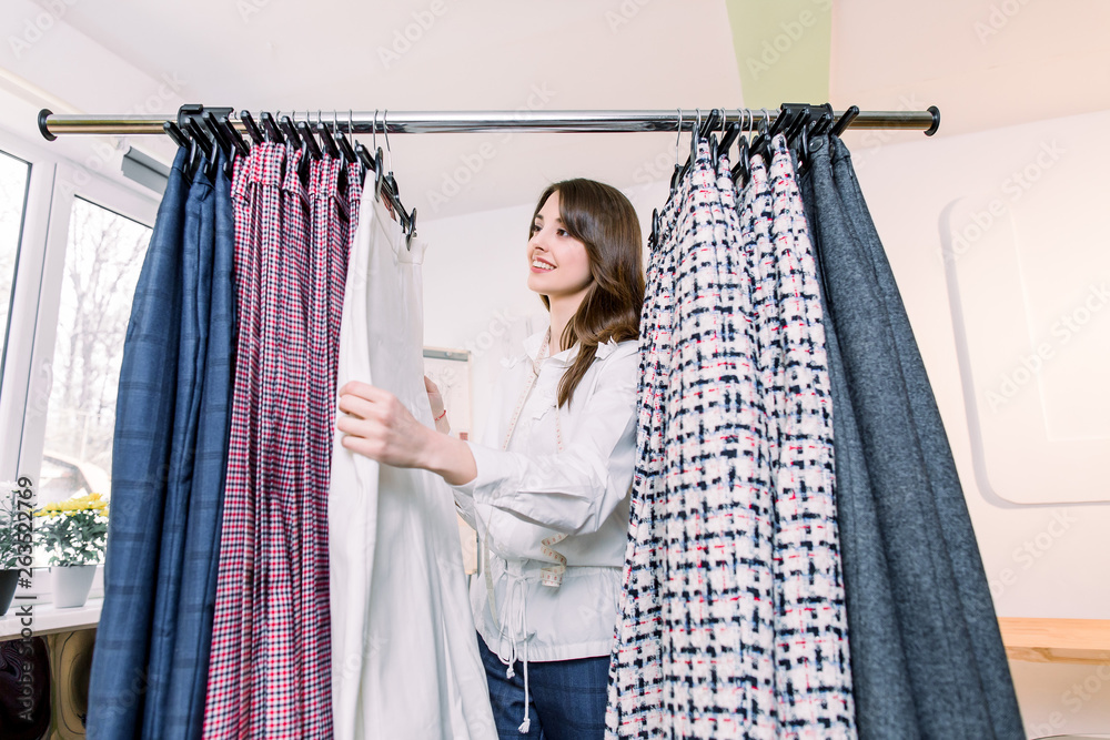 Pretty young Caucasian female designer or tailor standing near clothes rack with stylish pants, working in her atelier studio