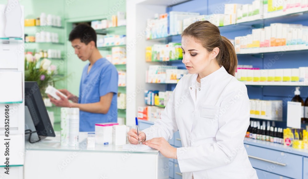 Young female pharmacist is attentively checking medicine with paper