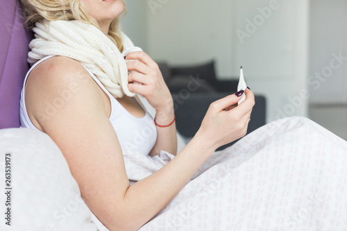Beautiful blond woman sitting in bed with a thermometer and a scarf wrapped around the neck and a handkerchief. illness and health at home