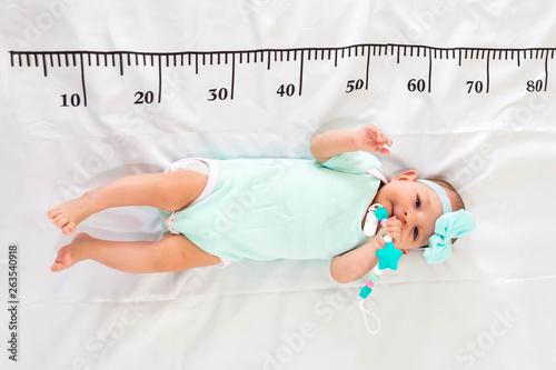 A three month baby in mint green clothes lying on a bed on which a measuring ruler for growth is drawn. Teething