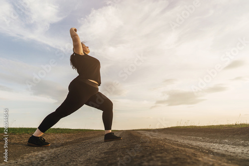 Young overweight woman doing morning yoga on the country road, open air activity. Healthy lifestyle, sport, weight loss