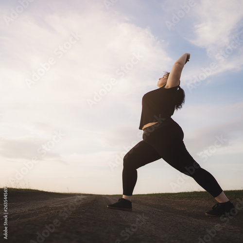 Young overweight woman doing morning yoga on the country road, open air activity. Healthy lifestyle, sport, weight loss