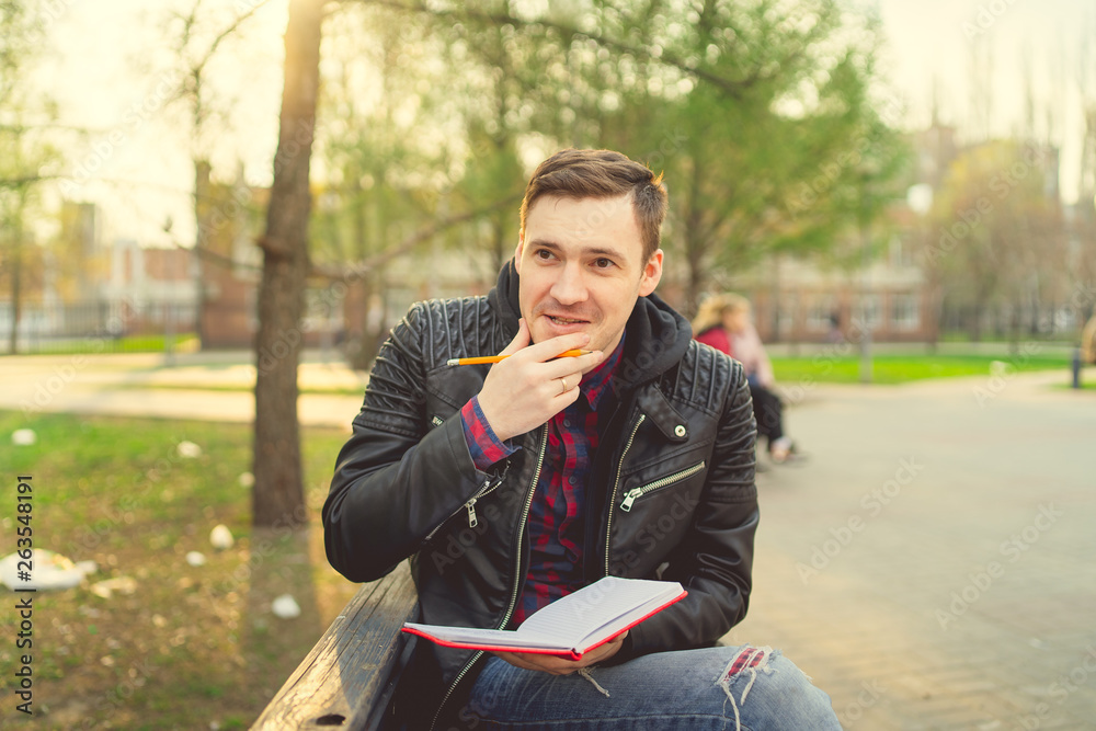 Man with a notebook in his hands in the Park. Male, student is writing her ideas and thoughts into the notebook sitting on the bench in the park Man with a notebook in his hands in the Park. Male, student is writing her ideas and thoughts into the notebook sitting on the bench in the park