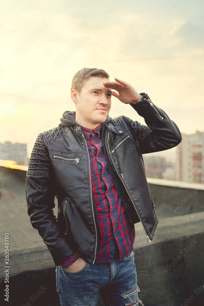 Man in trendy jacket on roof edge. Portrait of young male in leather jacket, standing on the roof of a multi-storey building. Man 30 years old at sunset Man in trendy jacket on roof edge. Portrait of young male in leather jacket, standing on the roof of a multi-storey building. Man 30 years old at sunset