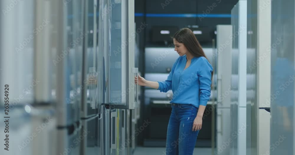 brunette woman in shirt to open the door of the refrigerator in the appliances store and compare with other models to buy the new house brunette woman in shirt to open the door of the refrigerator in the appliances store and compare with other models to buy the new house