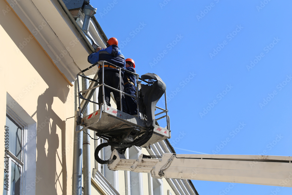 Lifting platform with workers against the blue sky, builders are repairing the roof gutter. Lifting crane basket for construction and repair works on background of building wall