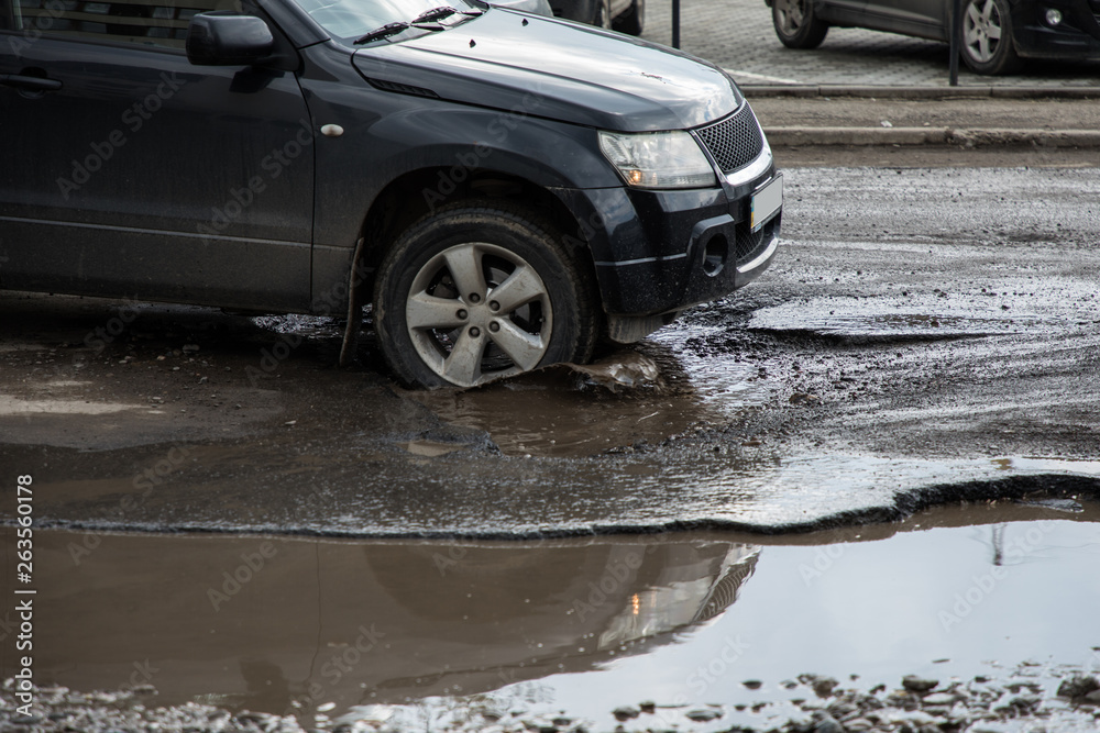 The car goes without reducing speed on a bad road with asphalt in pits and potholes, hitting the wheel in a puddle, spraying melt water in all directions