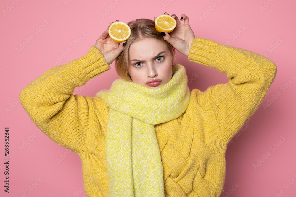 Human emotions. Blonde beautiful european girl in yellow sweater and scarf showing funny face, holding two sliced citrus lemons on her head Human emotions. Blonde beautiful european girl in yellow sweater and scarf showing funny face, holding two sliced citrus lemons on her head
