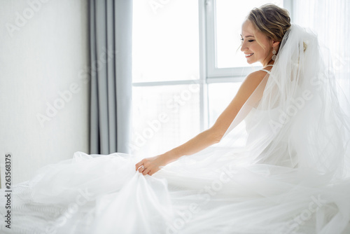 Amazing young bride posing in a beautiful wedding dress with wedding veil near window and smiling