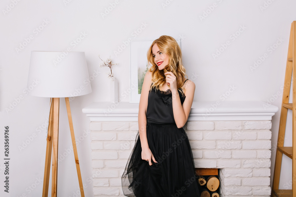 Portrait of young woman, blonde girl in bright room, studio with nice, modern white interion, standing next to fake fireplace, looking to side. Wearing stylish black dress. Portrait of young woman, blonde girl in bright room, studio with nice, modern white interion, standing next to fake fireplace, looking to side. Wearing stylish black dress.