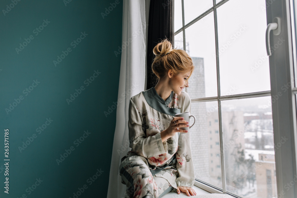 Young blonde woman drinking tea, coffee and looking through big window, happy, good morning at home. Wearing silk pajamas with flowers. Turquoise wall on background. - obrazy, fototapety, plakaty 