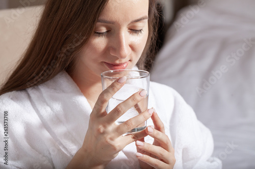 Young pretty european woman holding a glass and drink pure water sitting on the sofa. Cleansing the body of toxins. Healthy skin. Oily skin. Close up partrait of beautiful woman in bathrobe.