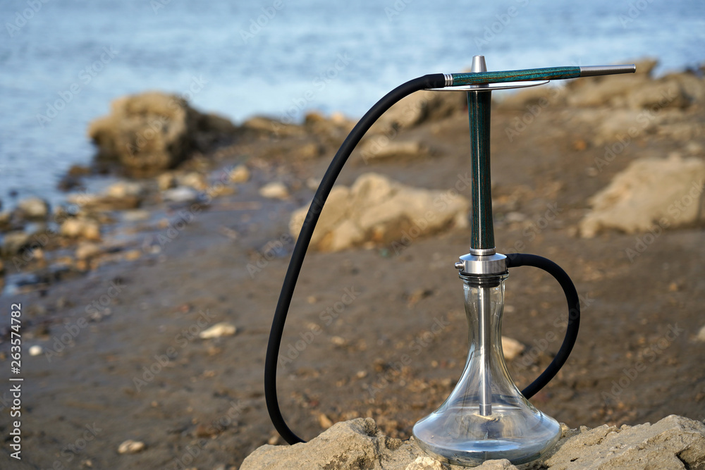 Blue wooden hookah with a black hose stands on the sea on a stone. Mouthpiece on a metal plate. Travel and concept for smoking hookah. Blurred background of nature and blue water.