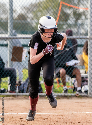 Female softball base runner smiling through her face mask while sprinting to first base.