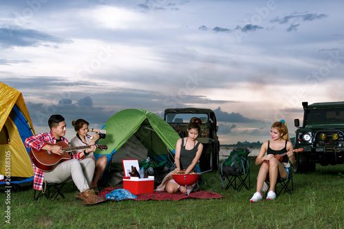 A group of Asian men and women are camping near a reservoir to relax and party. © nitinai2518