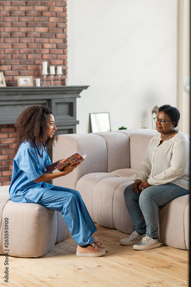 Dark-skinned private nurse talking to aged lady sitting on sofa