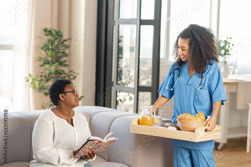 Caring nurse bringing tray with breakfast for aged woman