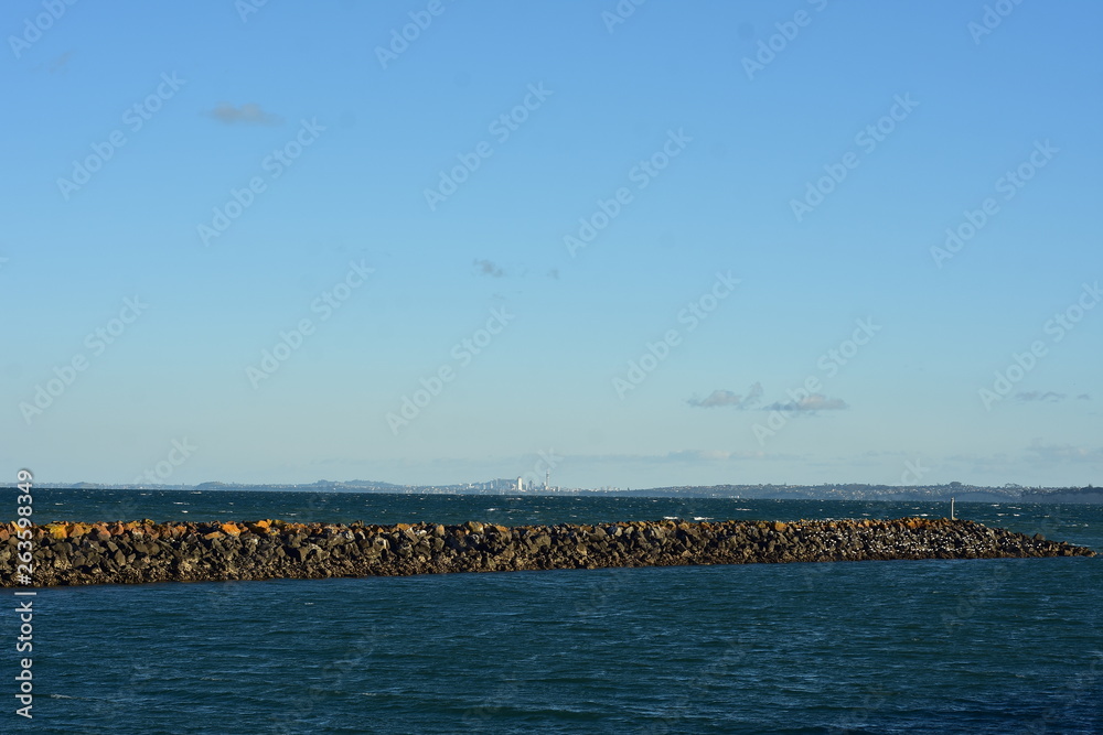 Fototapeta premium Breakwall of free laid stones surrounded by dark choppy sea with Auckland cityscape in distant background.