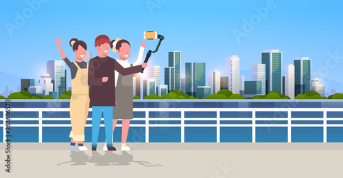 casual people using 3-axis gimbal stabilizer selfie stick for smartphone happy tourists taking photo standing together over cityscape background horizontal full length