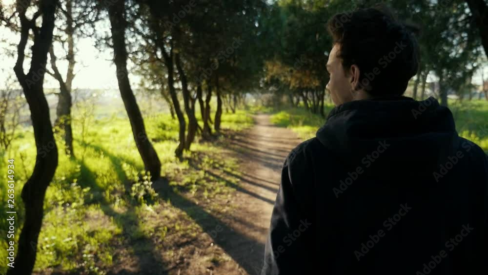 Young guy with curly hair walkng in the beautiful park on sunset. Green grass and trees trunks on background. Slow motion Young guy with curly hair walkng in the beautiful park on sunset. Green grass and trees trunks on background. Slow motion