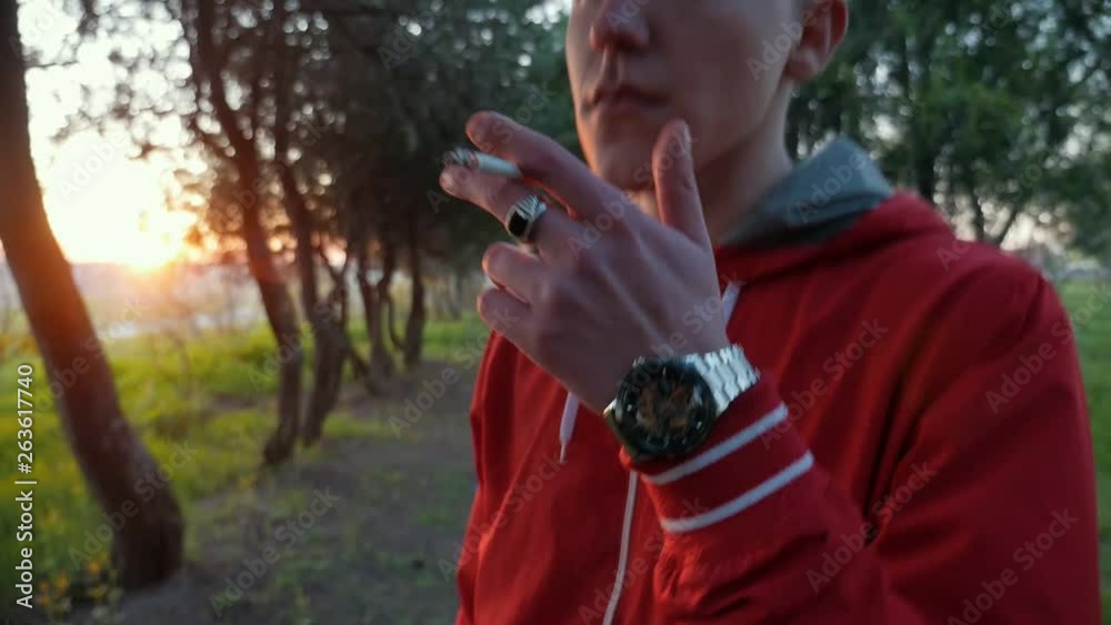 Camera follows the hand with cigarette of handsome young man smoking in the park at sunset. Face of smoking guy in slow motion