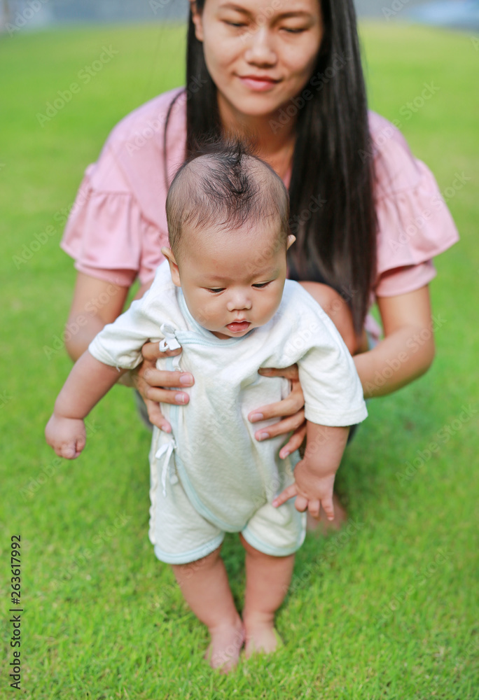 Fototapeta premium Mother training her infant baby to walk first steps on the green grass garden.