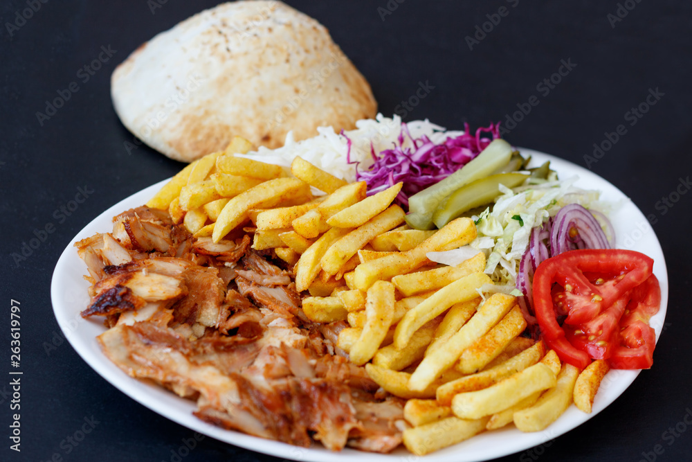 Chiken Doner Kebab on the plate with bread, french fries, tomatoes, onion, pickles and salad on a dark background. Grilled chicken meat with vegetables. Chiken Doner Kebab on the plate with bread, french fries, tomatoes, onion, pickles and salad on a dark background. Grilled chicken meat with vegetables.