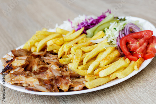 Chiken Doner Kebab on the plate with french fries, tomatoes, onion, pickles and salad on a wooden background. Grilled chicken meat with vegetables.