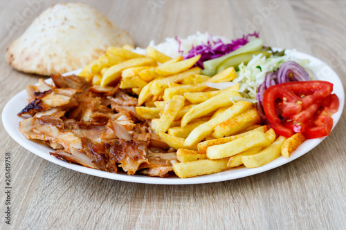 Chiken Doner Kebab on the plate with bread, french fries, tomatoes, onion, pickles and salad on a wooden background. Grilled chicken meat with vegetables.