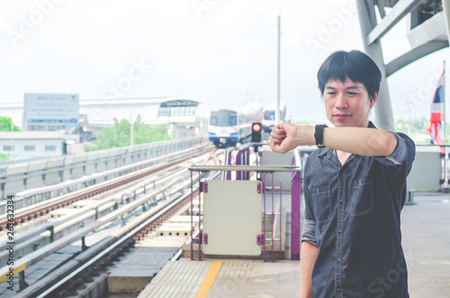 View of asian man stand and looking at his watch while waiting for something or worrying about time. Motion train as background. Business and travel concept.