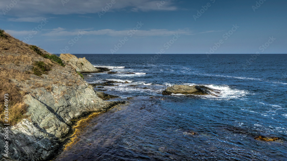 Rocky cliff shore by a seaside, small waves, green horizon, blue sky with some smooth clouds