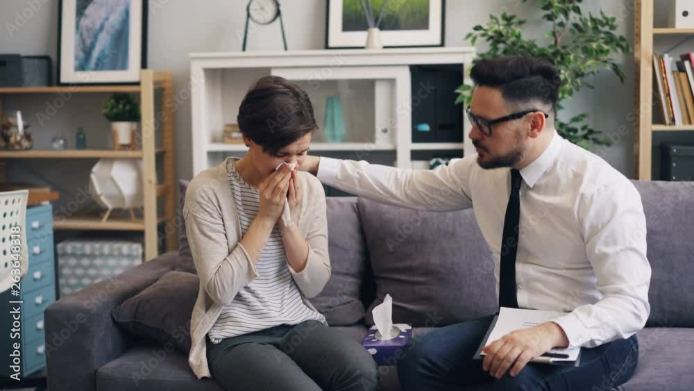 Helpful male therapist is comforting stressed woman crying in office giving her paper tissue giving advice sitting on couch together having conversation discussing problem. Helpful male therapist is comforting stressed woman crying in office giving her paper tissue giving advice sitting on couch together having conversation discussing problem.