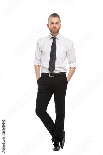 A young man with a beard in his daily shirt stands tall and holds his hands in his trouser pockets, isolated on a white background.