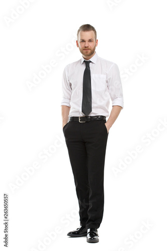 A young man with a beard in his daily shirt stands tall and holds his hands in his trouser pockets, isolated on a white background.