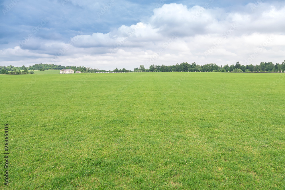 Rural landscape. The flat well-groomed field of a short-haired grass, a country house in the distance at the hill. The beautiful cloudy sky in sunny day