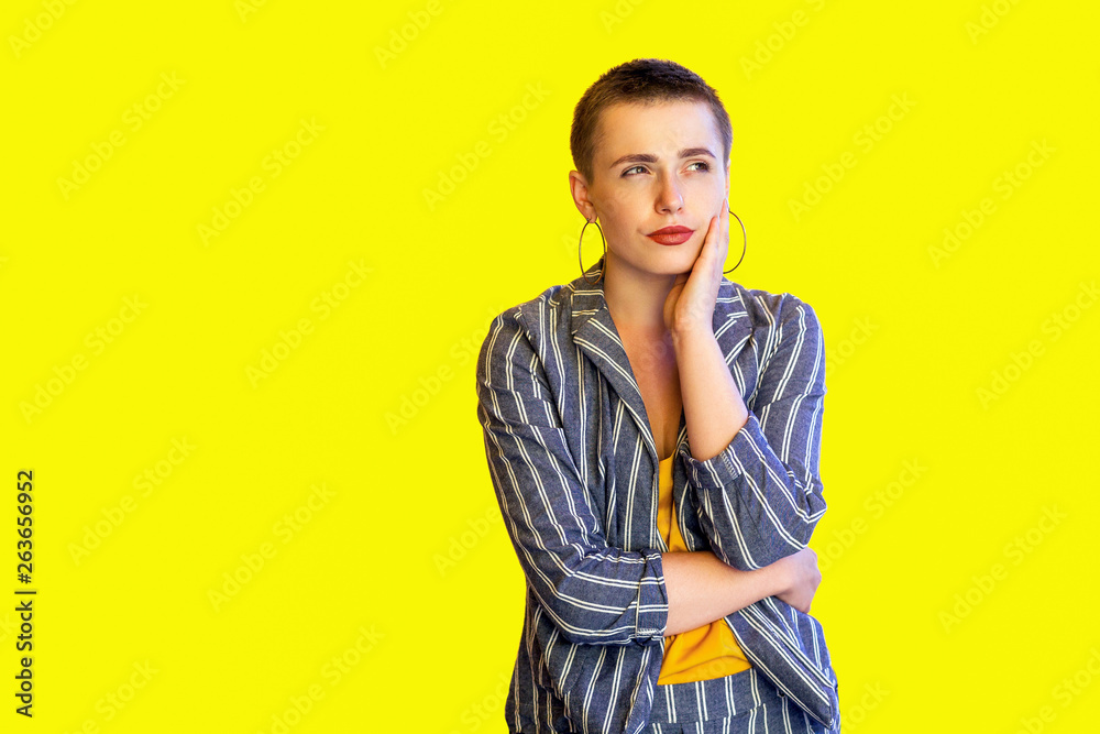 Portrait of thoughtful young short hair fancy casual stylish woman in yellow shirt and striped suit standing touching her face and thinking what to do. indoor studio shot isolated on yellow background Portrait of thoughtful young short hair fancy casual stylish woman in yellow shirt and striped suit standing touching her face and thinking what to do. indoor studio shot isolated on yellow background