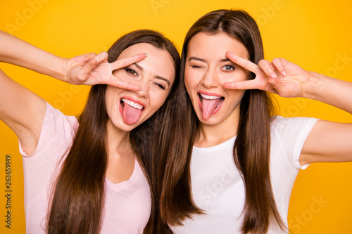 Close up photo of cute funny pretty girls trendy students best have free time travel make v-signs near eyes tongue-out feel satisfied careless isolated wear light-colored t-shirts yellow background