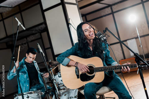 selective focus of beautiful inspired musician playing guitar and singing while mixed race man playing drums in recording studio