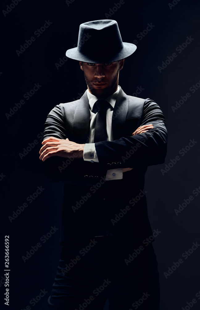 Handsome confident man in black suit and hat with arms crossed on dark background Handsome confident man in black suit and hat with arms crossed on dark background