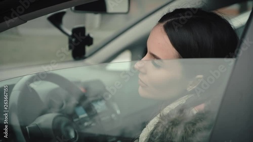 A beautiful young girl sits behind the wheel of her car and raises the window of the door.