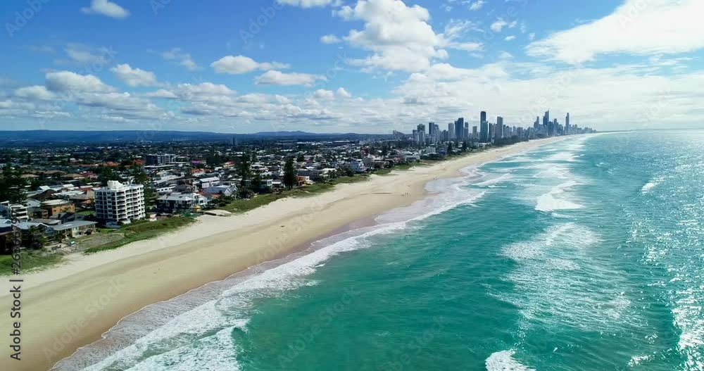 Sliding drone shot of the Mermaid Beach, Queensland, Australia, with the sandy beach between the coastline and the city, with skyscrapers on the background, on a bright sunny day with some clouds