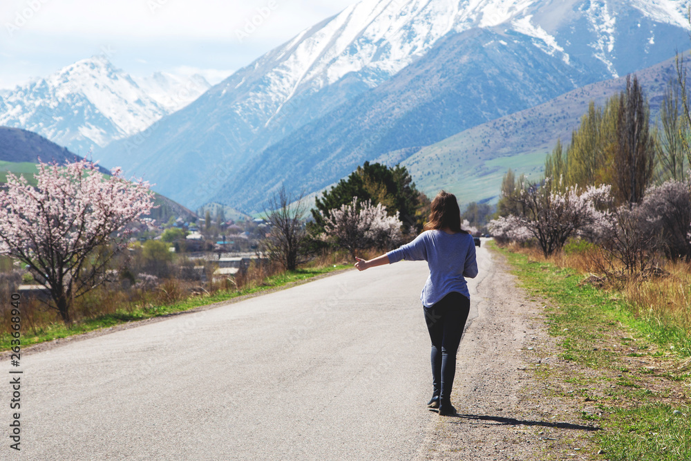 A young girl on the background of a mountain road catches the car. Spring portrait outside the city. Flowering trees and mountains. Recreation and adventure.