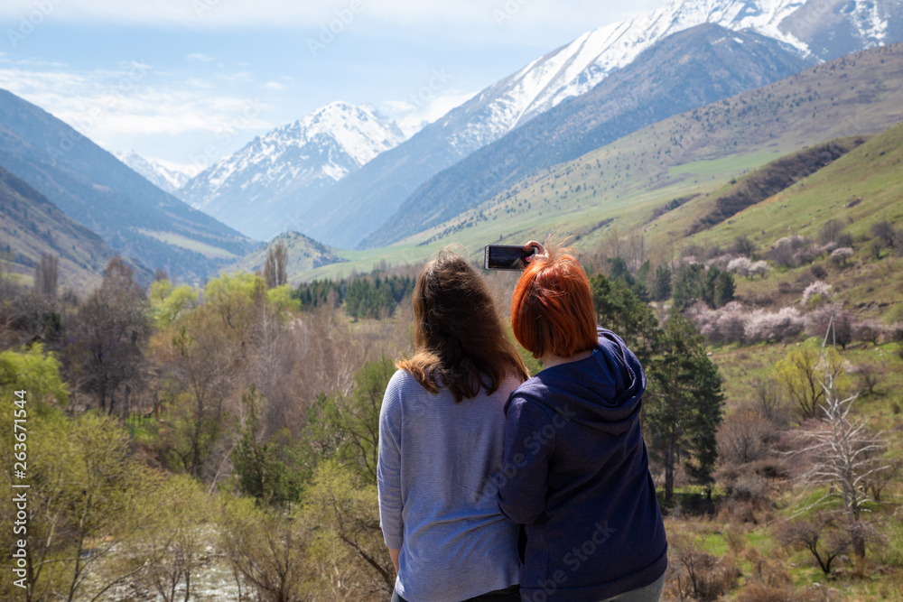 Two girls happily photographed against the backdrop of the mountains. Travelers take a selfie on the background of a beautiful mountain gorge. High snowy mountains and green trees in front of them.