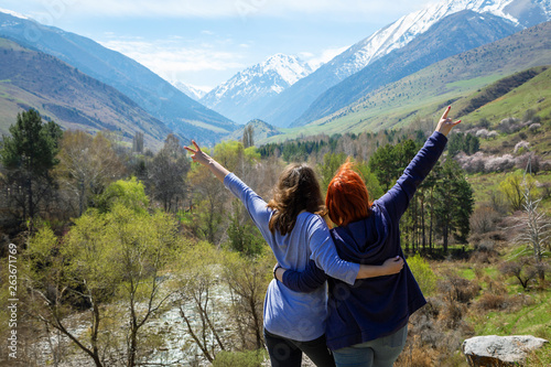 Two girls happily raise their hands up. Travelers on the background of a beautiful mountain gorge. High snowy mountains and green trees in front of them.
